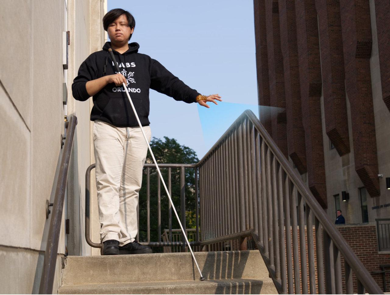 A blind person is walking downstairs outdoors, using a white cane. They use the hand device to look for handrails to hold on.